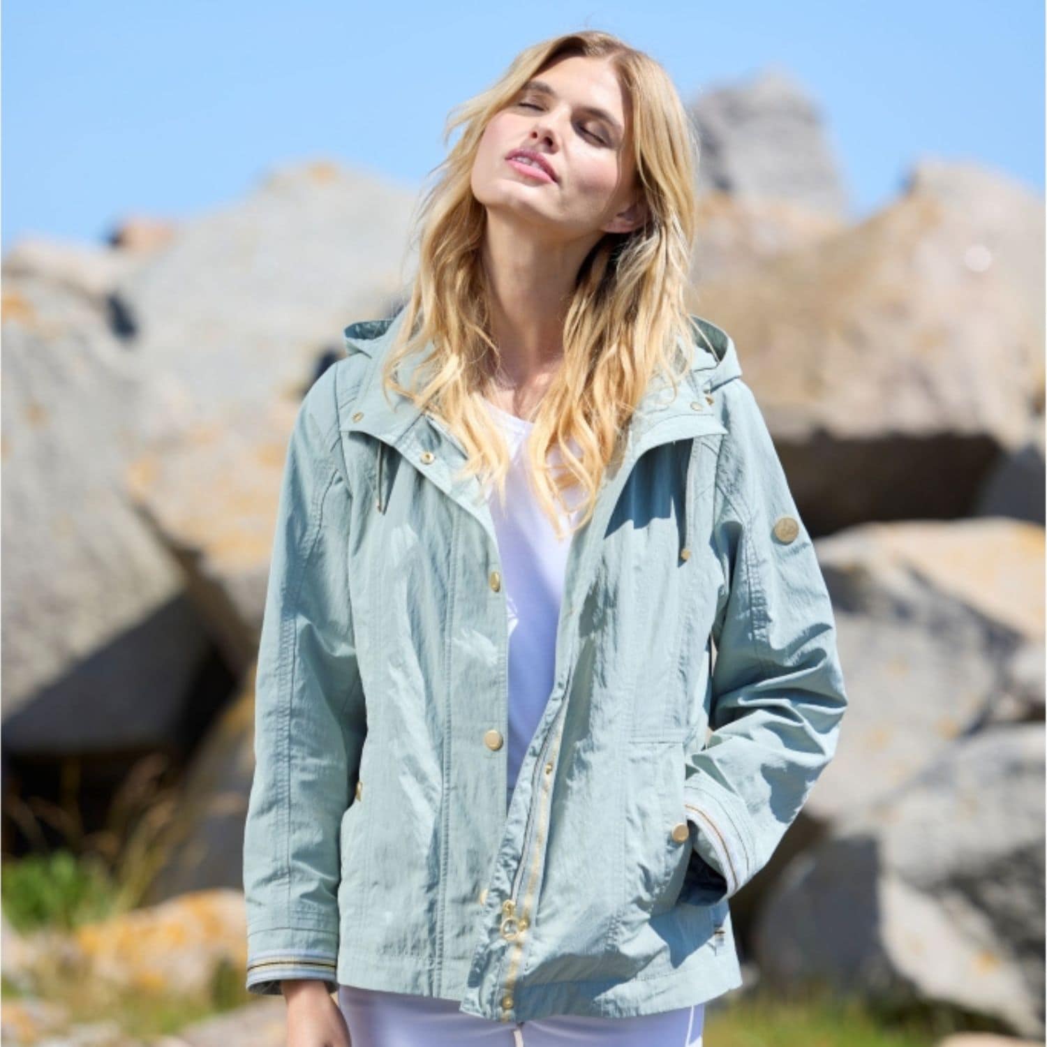 Lightweight summer jacket for women in sage with gold buttons and striped cuff details in front of rocks in summer sun.